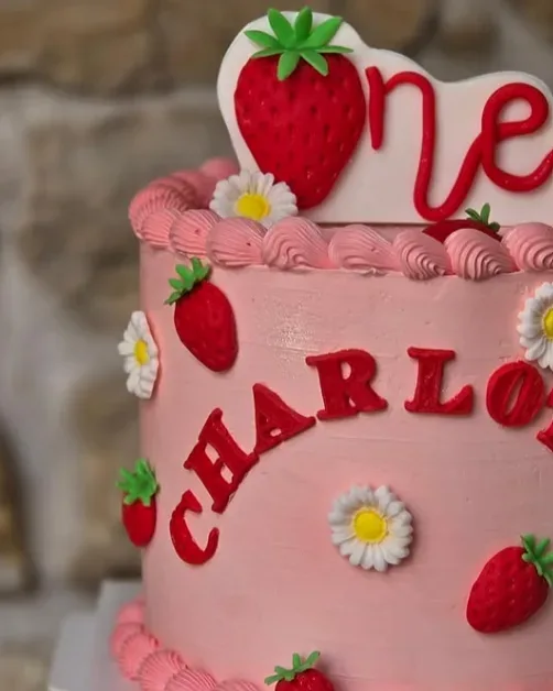 Gros plan sur un gâteau rose de premier anniversaire avec lettres rouges Charlotte, fraises modelées, marguerites et pochage régulier